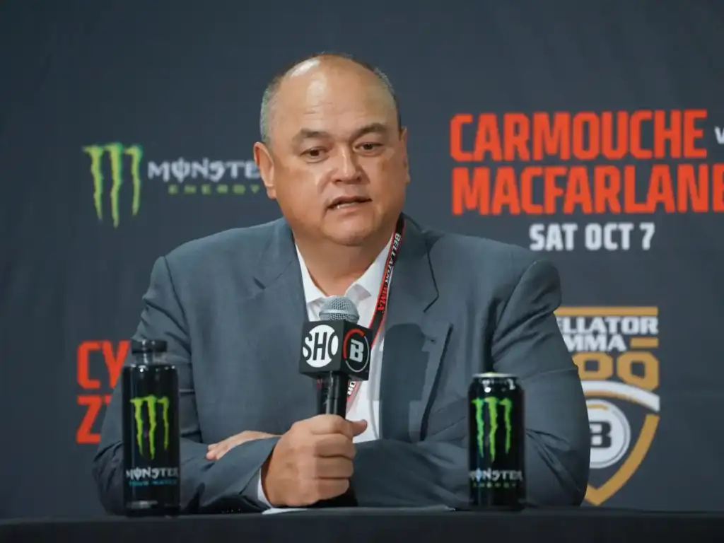 Man in a suit speaks into a microphone at a press conference, with Monster energy cans on the table.