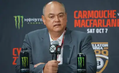 Man in a suit speaks into a microphone at a press conference, with Monster energy cans on the table.