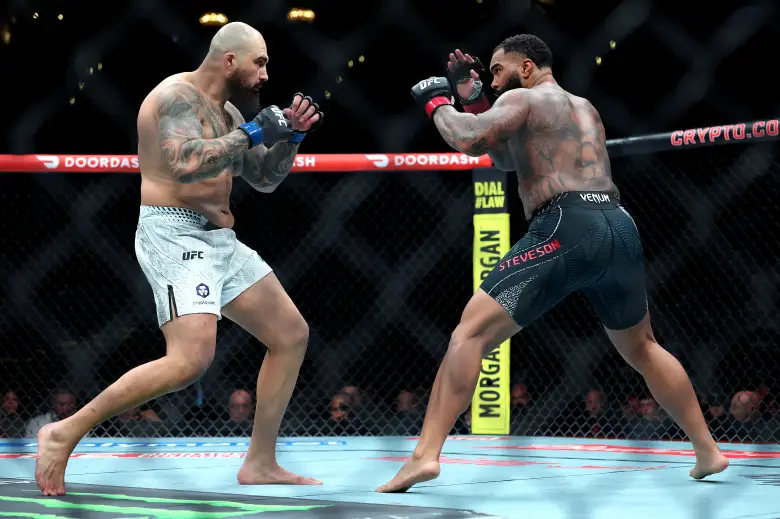Two UFC fighters wearing gloves face off in the cage during a match, one in white shorts, the other in dark shorts, crowd in background.