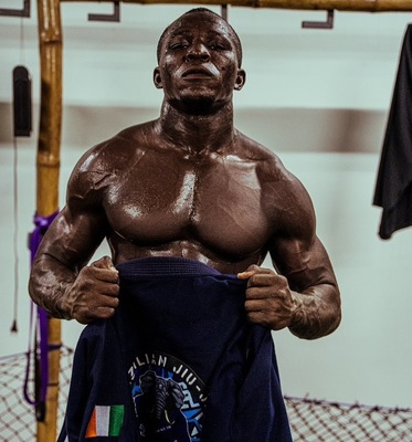 Muscular man in a gym pulling up a shirt, revealing a graphic tee and a determined expression between sweat and lighting.