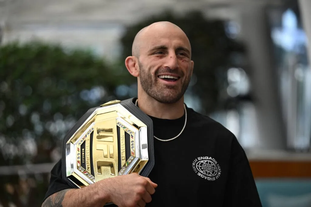 0f9e5529e43c2f11b3f816182cb86e8d - AfroCombat Man proudly displaying a UFC championship belt over his shoulder while smiling.
