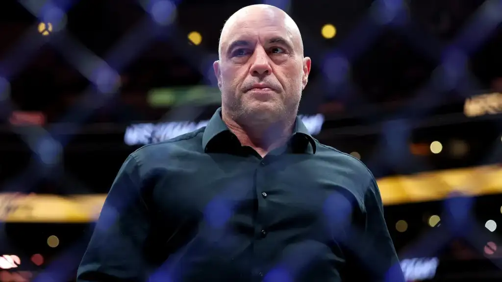 Bald man in a dark button-down shirt stands inside a fenced arena, lights blurred in the background at a sports event.