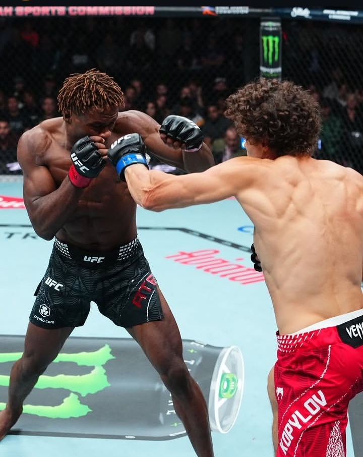 Two UFC fighters in the octagon exchange punches; one wears blue gloves and black shorts, the other wears red shorts, with a cheering crowd in the background.