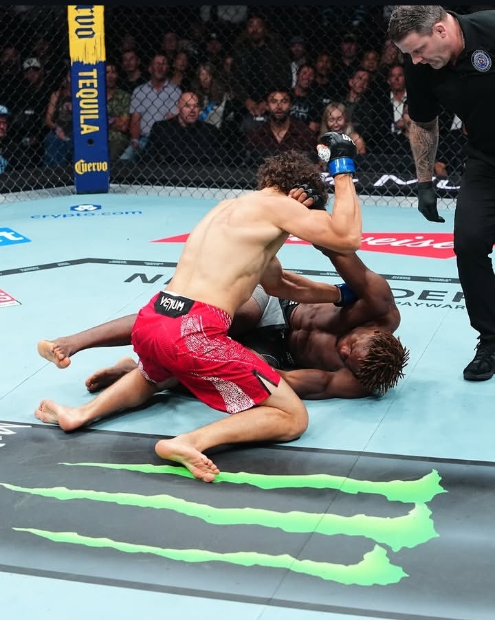 Two shirtless mixed martial artists grapple on a blue octagon floor while a referee watches, with a crowd of spectators behind the cage.