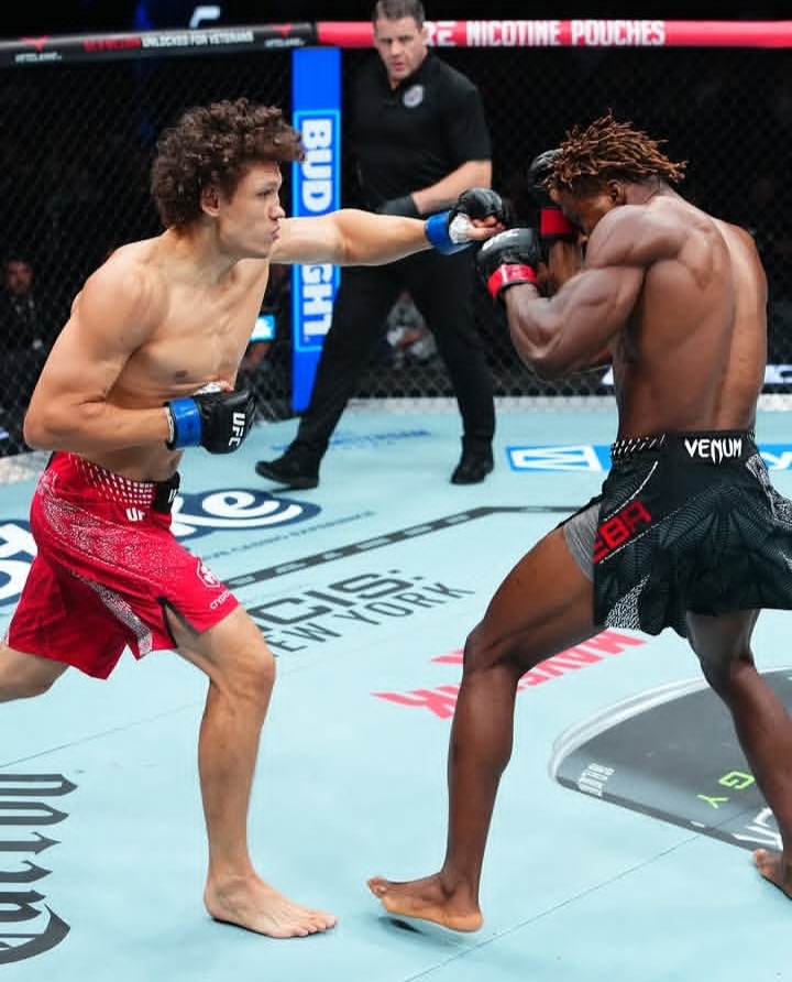 Two mixed martial artists exchange punches inside a UFC octagon, one delivering a left jab while the other blocks with his guard; referee in the background.