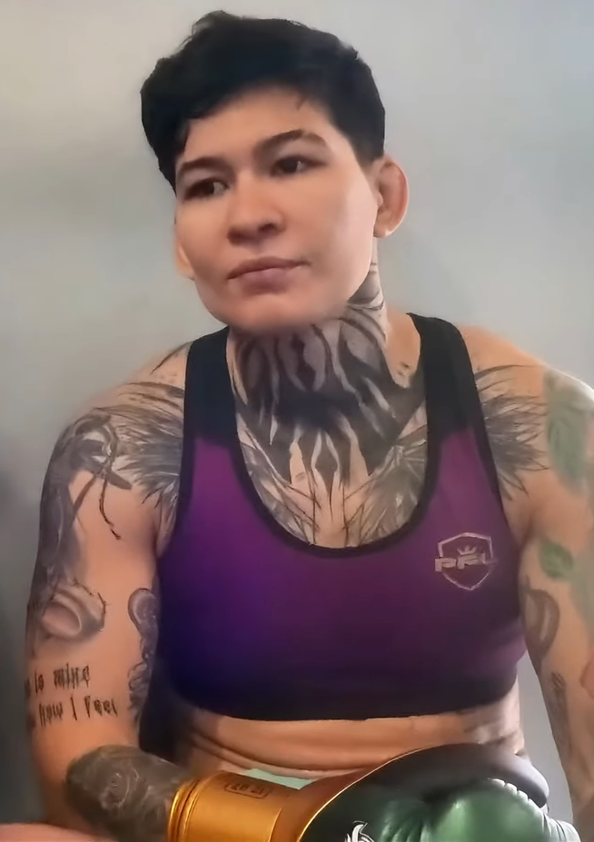 Person with short black hair wearing a purple sports bra, heavily tattooed neck and arms, sitting with green boxing gloves on