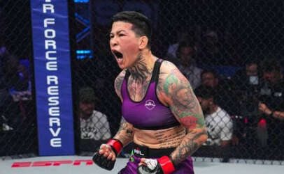 Female MMA fighter in a purple sports bra and shorts, shouting with fists raised inside the cage. She has visible tattoos and fans watch from the stands.