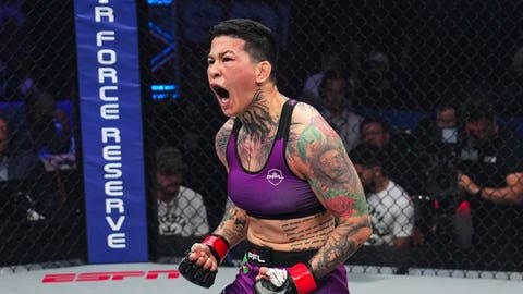 Female MMA fighter in a purple sports bra and shorts, shouting with fists raised inside the cage. She has visible tattoos and fans watch from the stands.
