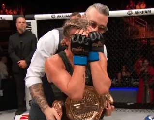 Female MMA fighter with blue gloves sits in the octagon, holding a championship belt, as a man hugs her from behind in celebration.