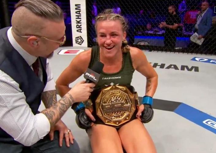 Female MMA fighter sits in the cage after winning, smiling while holding a gold championship belt as a reporter interviews her.