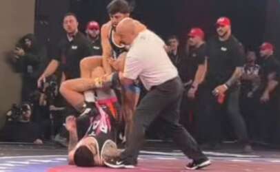 Wrestling match scene: a referee in a white shirt pins a grappler on a red mat while another competitor watches, with audience and photographers in the background.