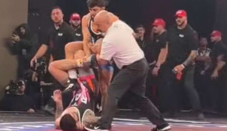 Wrestling match scene: a referee in a white shirt pins a grappler on a red mat while another competitor watches, with audience and photographers in the background.