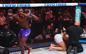 MMA fight inside the cage; a fighter in purple shorts charges toward an opponent on the mat as the referee kneels nearby, with the crowd watching.