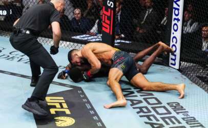 MMA fight on the canvas: one fighter pins the other on the mat inside a UFC-branded cage as a referee watches nearby.