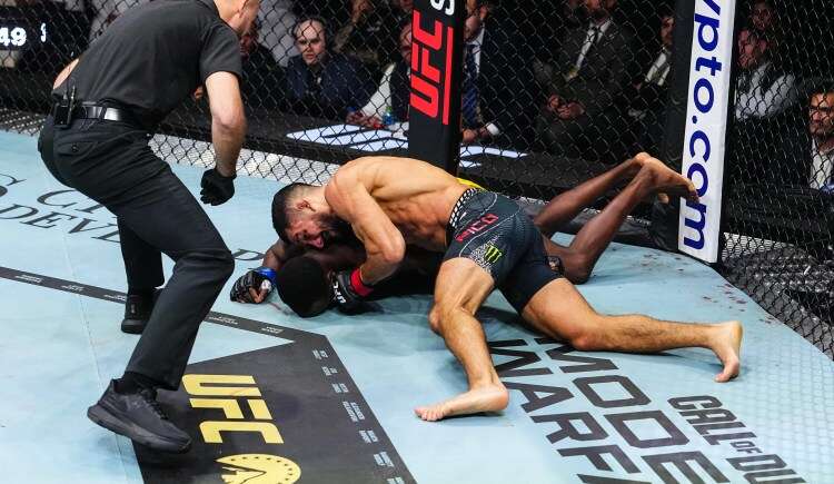 MMA fight on the canvas: one fighter pins the other on the mat inside a UFC-branded cage as a referee watches nearby.