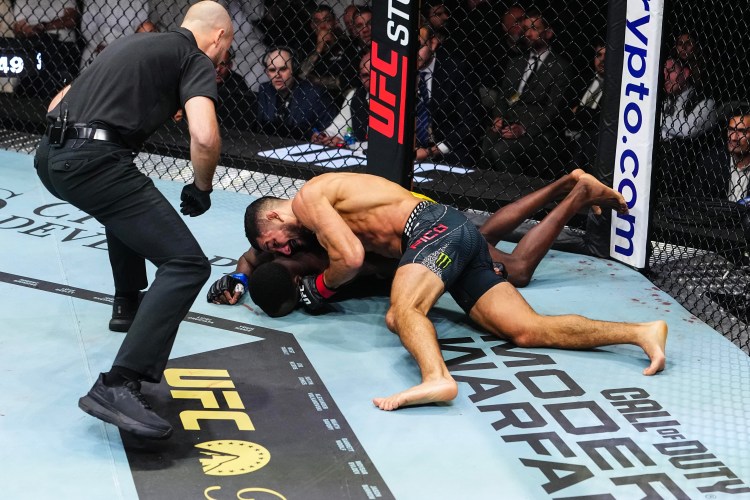 MMA fight on the canvas: one fighter pins the other on the mat inside a UFC-branded cage as a referee watches nearby.