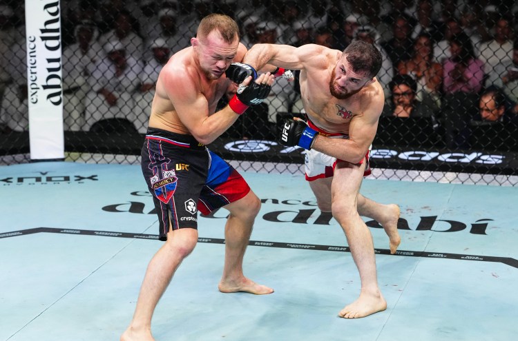 Two shirtless UFC fighters grapple in the cage; one throws a punch while the other blocks, surrounded by spectators.