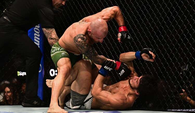 Two MMA fighters clash inside the cage; one man standing over the other delivering punches as the grounded opponent defends with his arms raised. A referee watches from the left.
