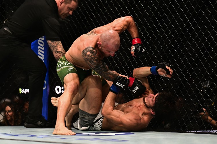 Two MMA fighters clash inside the cage; one man standing over the other delivering punches as the grounded opponent defends with his arms raised. A referee watches from the left.