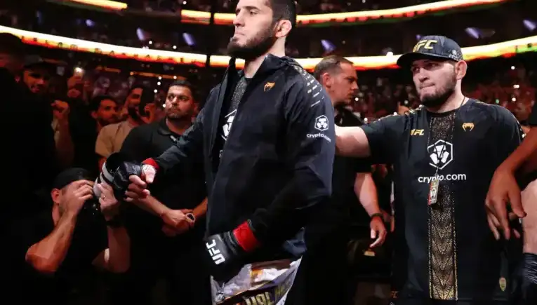 UFC fighter in a black jacket and gloves walks toward the octagon as crowd cheers, entourages nearby.