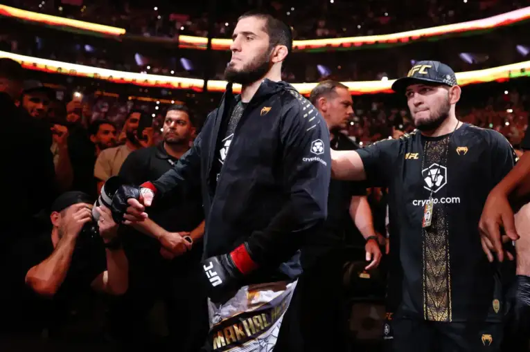 UFC fighter in a black jacket and gloves walks toward the octagon as crowd cheers, entourages nearby.