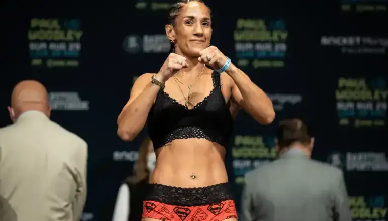 Female fighter in a black lace sports bra and red patterned shorts poses with fists up on a weigh‑in stage, with banners in the background.
