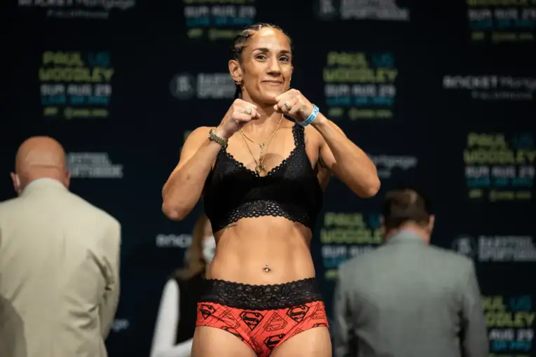 Female fighter in a black lace sports bra and red patterned shorts poses with fists up on a weigh‑in stage, with banners in the background.