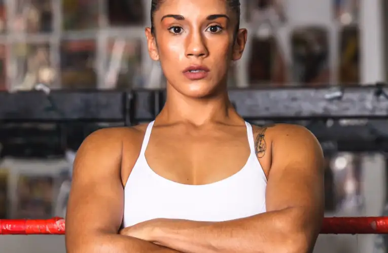 Athletic woman in a white sports top stands with arms crossed inside a gym boxing ring, looking ahead.