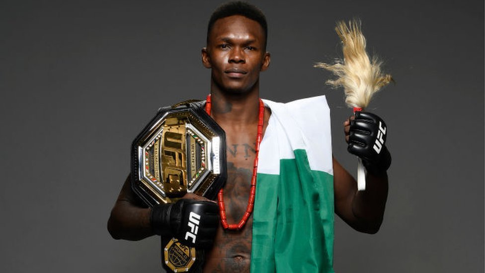 UFC champion posing with the championship belt on his shoulder/waist, wearing gloves and a Nigerian flag draped over his torso, holding a plume in one hand.