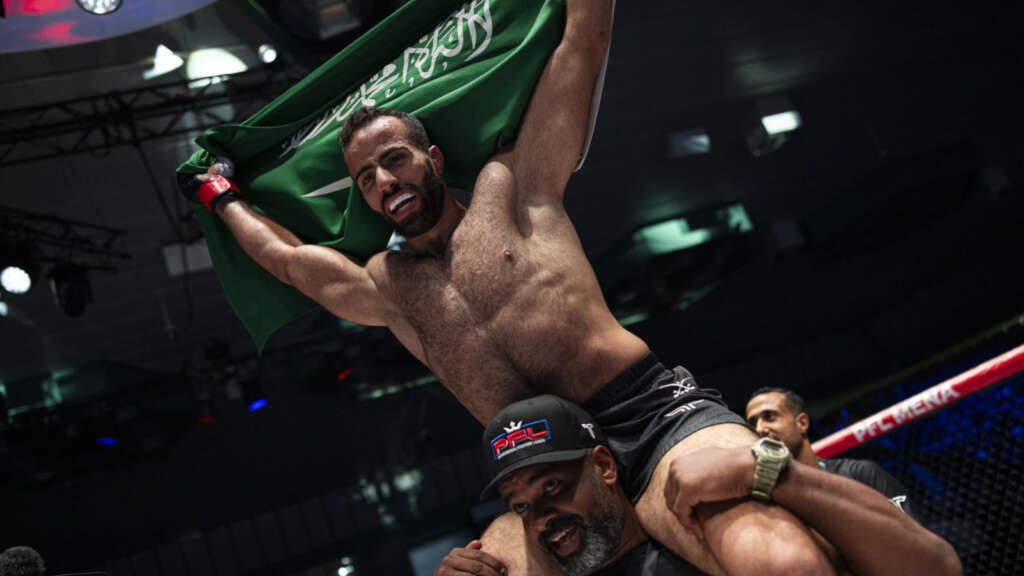 Shirtless fighter hoists a green Saudi Arabian flag above his head in a boxing ring, celebrating a win.