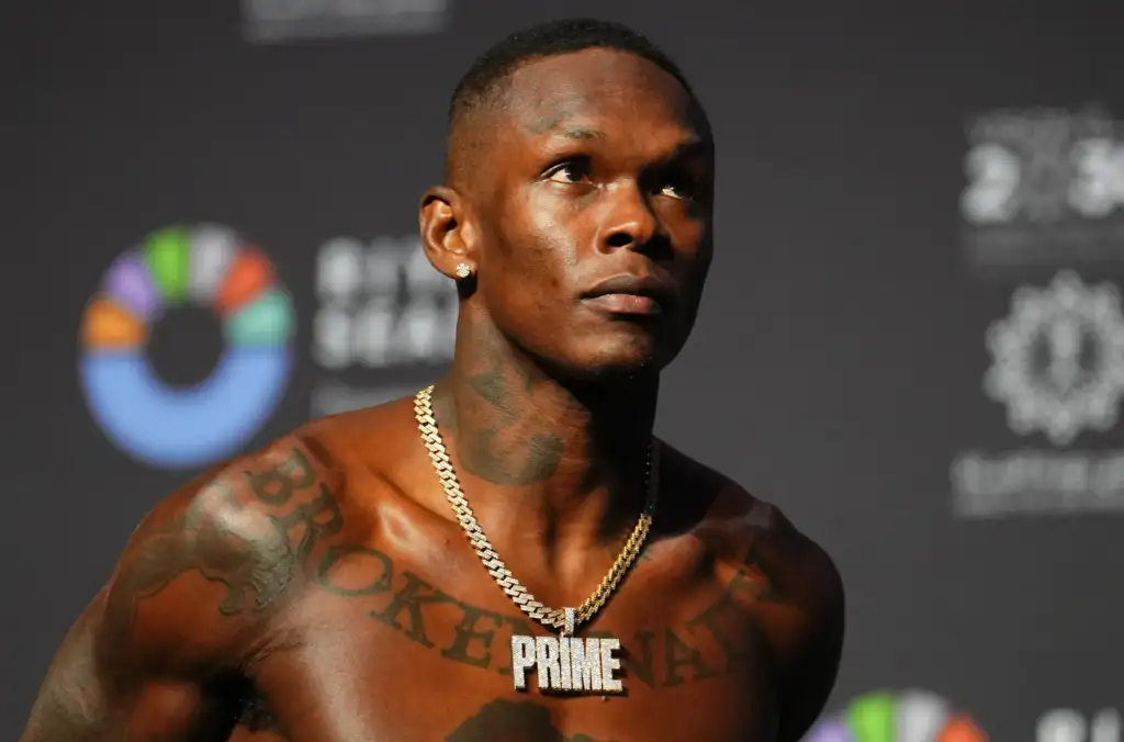 Shirtless man with tattoos and a gold chain, looking upward against a dark background with a blurred event banner behind him.