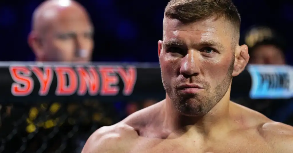 Close-up of a muscular male fighter's face in a cage, sweating, with a blurred crowd and a Sydney sign in the background.