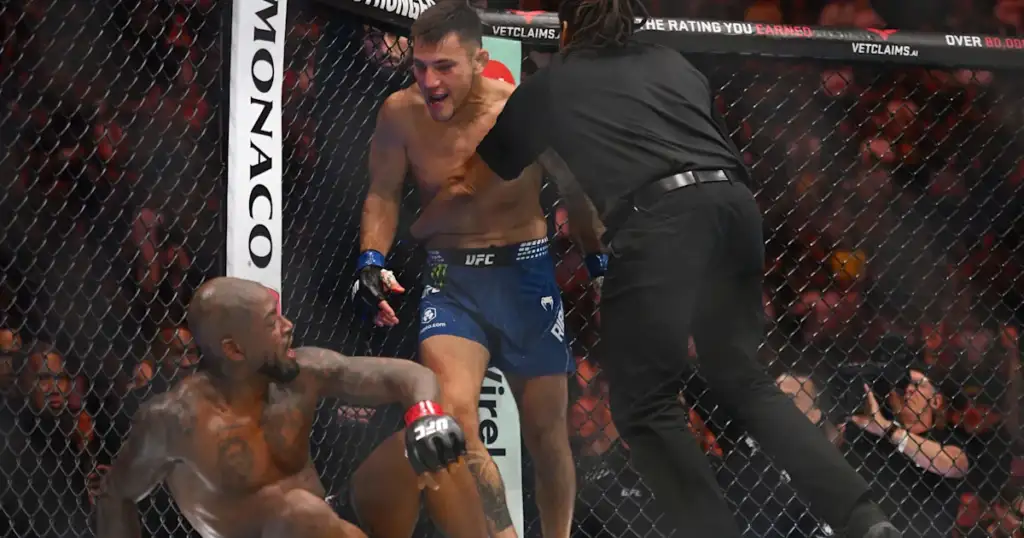 UFC match inside a cage: a fighter in blue shorts stands over a fallen opponent while a referee moves in nearby.