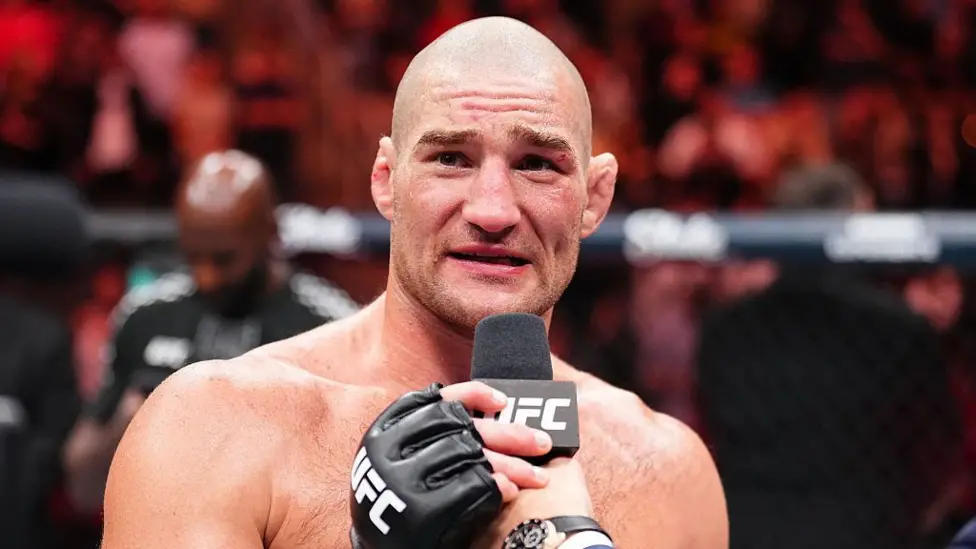 UFC fighter speaks into a microphone during a post-fight interview in the octagon, crowd blurred in the background.