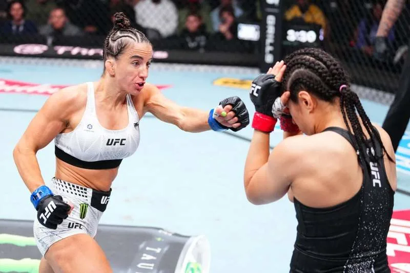 Two female UFC fighters clash in the octagon; the fighter in white throws a punch toward the opponent in black who shields with raised gloves in defense.