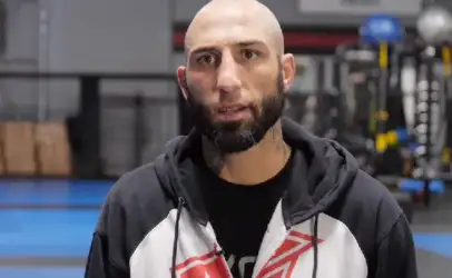 Bald man with a beard wearing a black and white hoodie speaks to the camera in a gym setting.