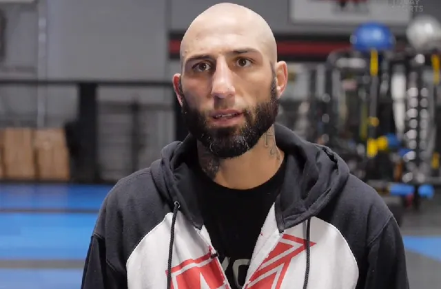 Bald man with a beard wearing a black and white hoodie speaks to the camera in a gym setting.