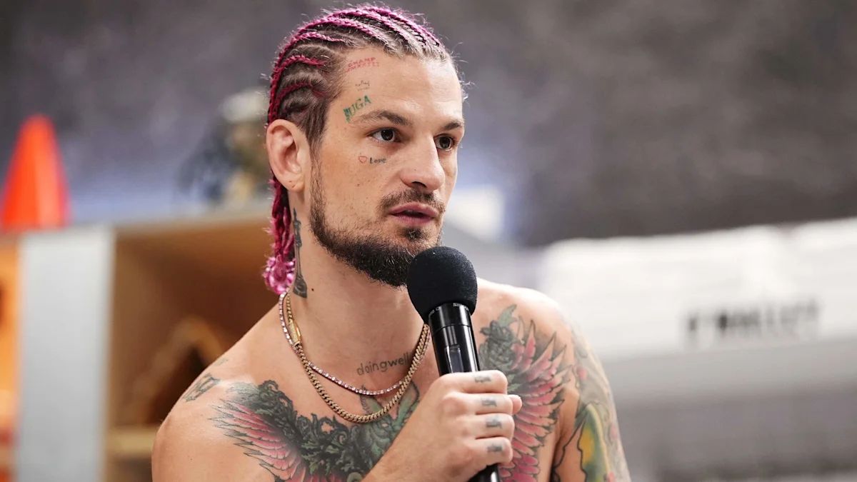 Tattooed man with pink cornrow hair speaks into a handheld microphone in a studio setting.