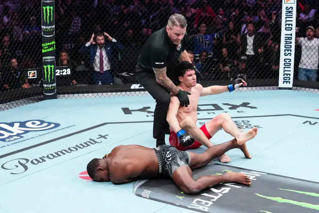 MMA referee restrains a downed fighter on the mat as a rival in red shorts sits nearby in the octagon, crowd watching in the background.