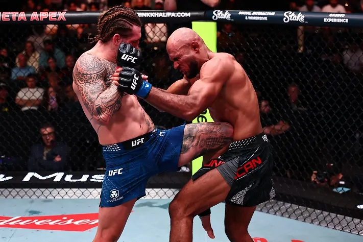 AllenDuncanjpg - AfroCombat Two UFC fighters in the octagon clash in a close clinch as one lifts a knee toward the other’s torso, both wearing gloves and shorts, with a crowd watching behind the cage.
