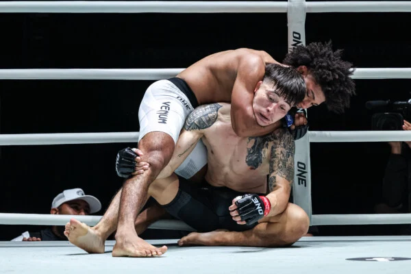 Two male mixed martial artists grapple on the mat inside a boxing/MMA ring, one in a top position applying a hold on the other.