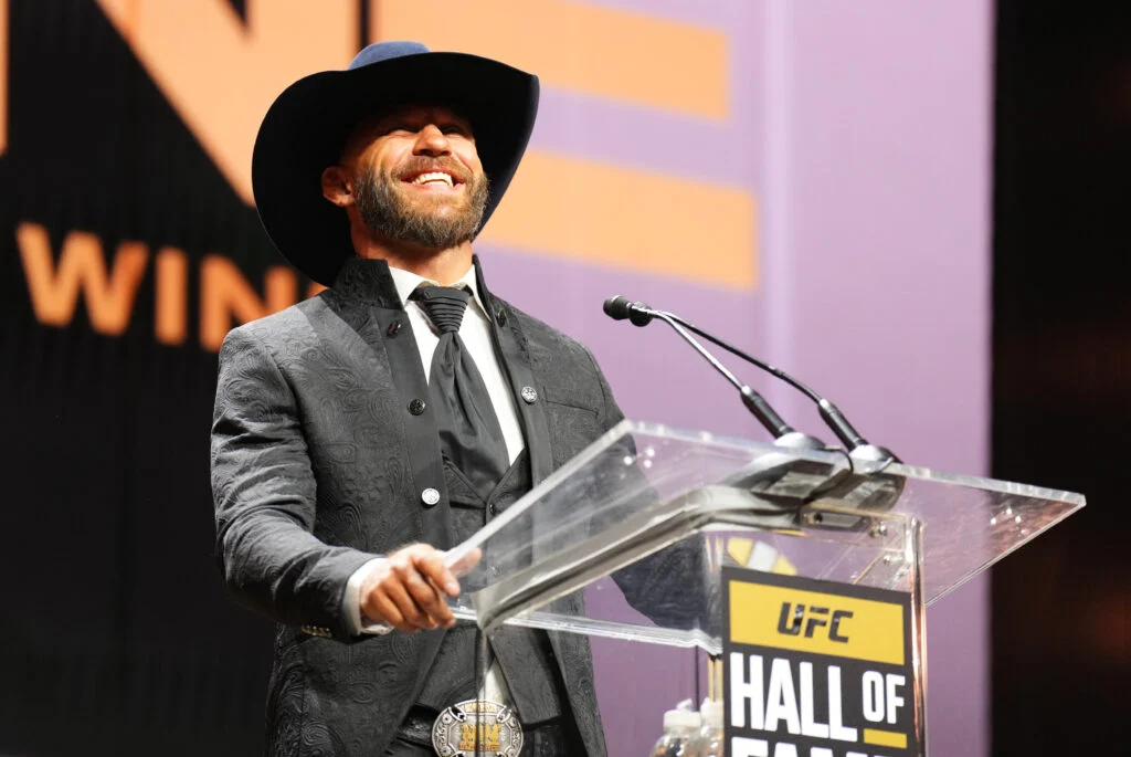 Man wearing a wide-brimmed black cowboy hat and patterned suit speaks at a UFC Hall of Fame podium.