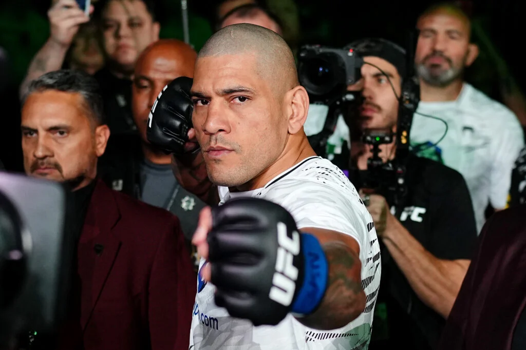 MMA fighter in white shirt throws a punch toward the camera as photographers and crowd surround him at an event.