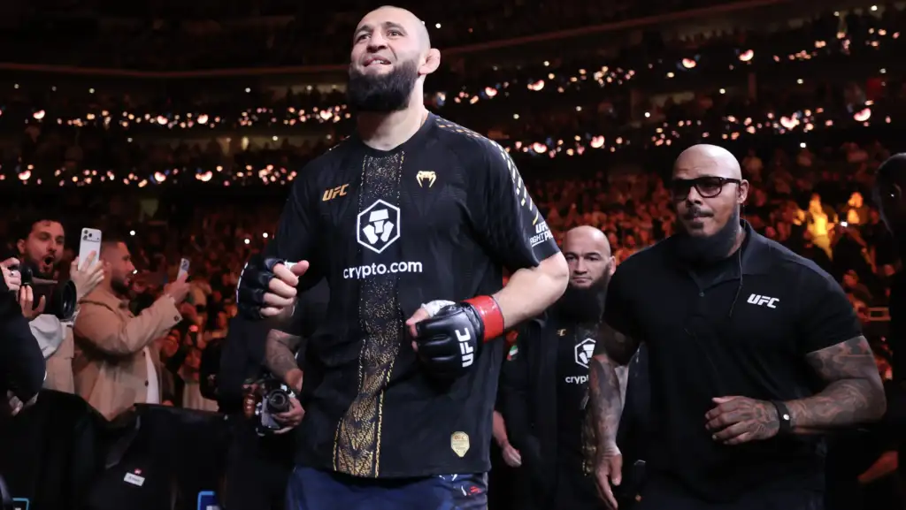 Bearded UFC fighter in a black and gold Crypto.com jersey walks toward the octagon as a large crowd cheers and cameras flash around him.