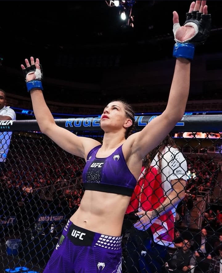 UFC fighter in purple gear raises both arms in victory inside the cage, crowd cheering behind.