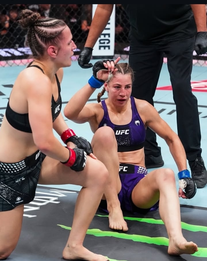 Two female UFC fighters on the mat inside the cage; one in black gear stands while the other, in purple, sits and gestures with braided hair as a referee stands nearby.