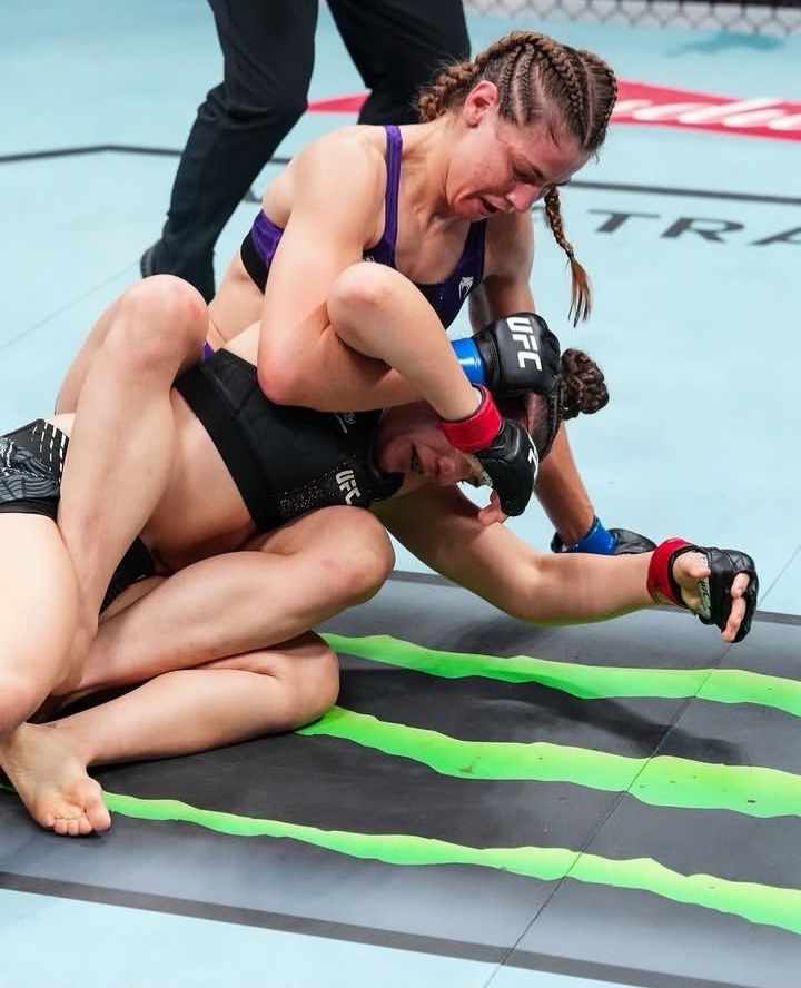 Female mixed martial arts fighters grapple on the mat inside the octagon during a match, one pinning the other from the side grip with gloves on.