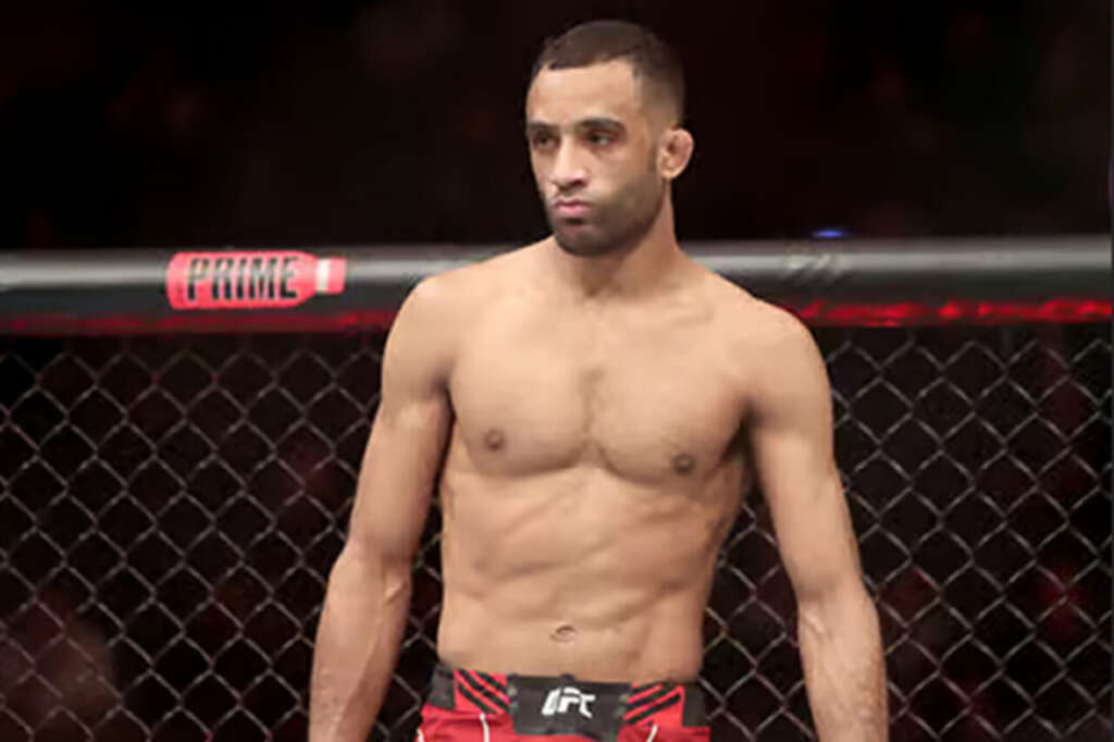 Male mixed martial artist standing shirtless inside a chain-link cage, wearing red shorts and looking to the side.