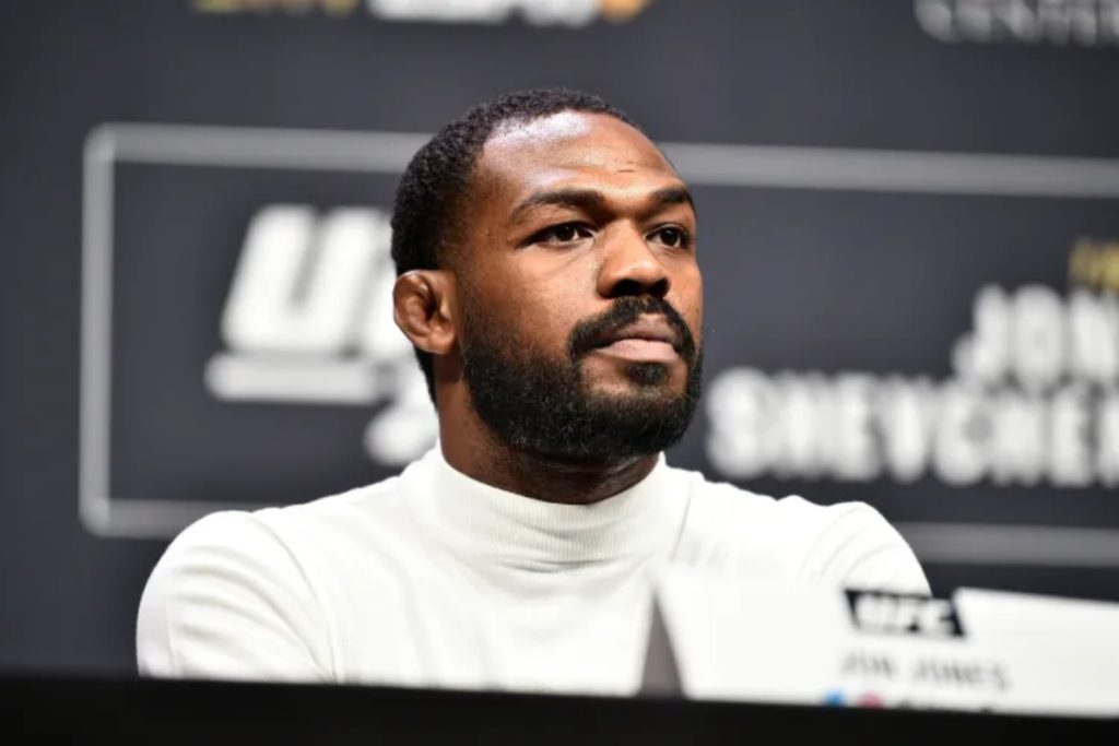 Close-up of a man with a beard in a white turtleneck at a UFC press conference, with branding behind him.