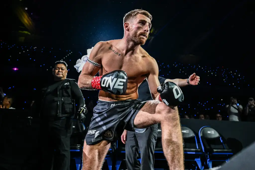 Male mixed martial artist in black shorts and red hand wraps delivering a kick in a ONE Championship ring with a cheering audience behind him.